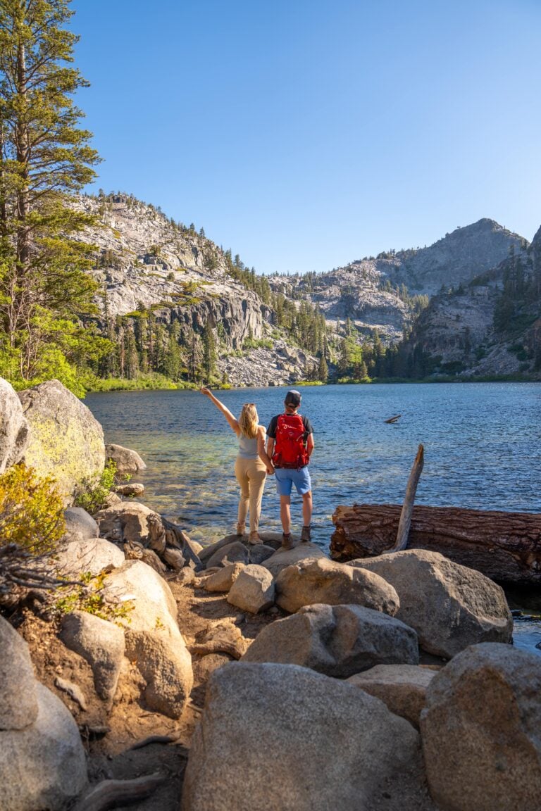 A couple stands on rocky ground at the edge of a clear mountain lake, surrounded by pine trees and mountains, with one person pointing toward the scenic view under a clear blue sky.