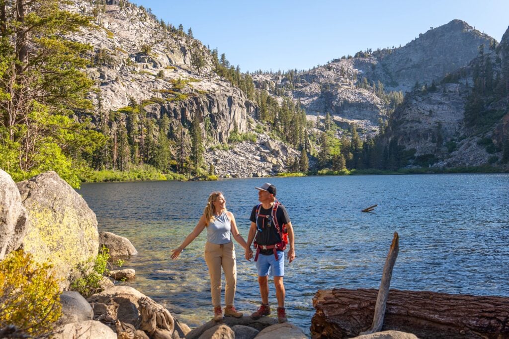 A couple stands on rocky ground by a clear mountain lake, surrounded by pine trees and rugged cliffs under a bright blue sky, enjoying a scenic outdoor hike together.