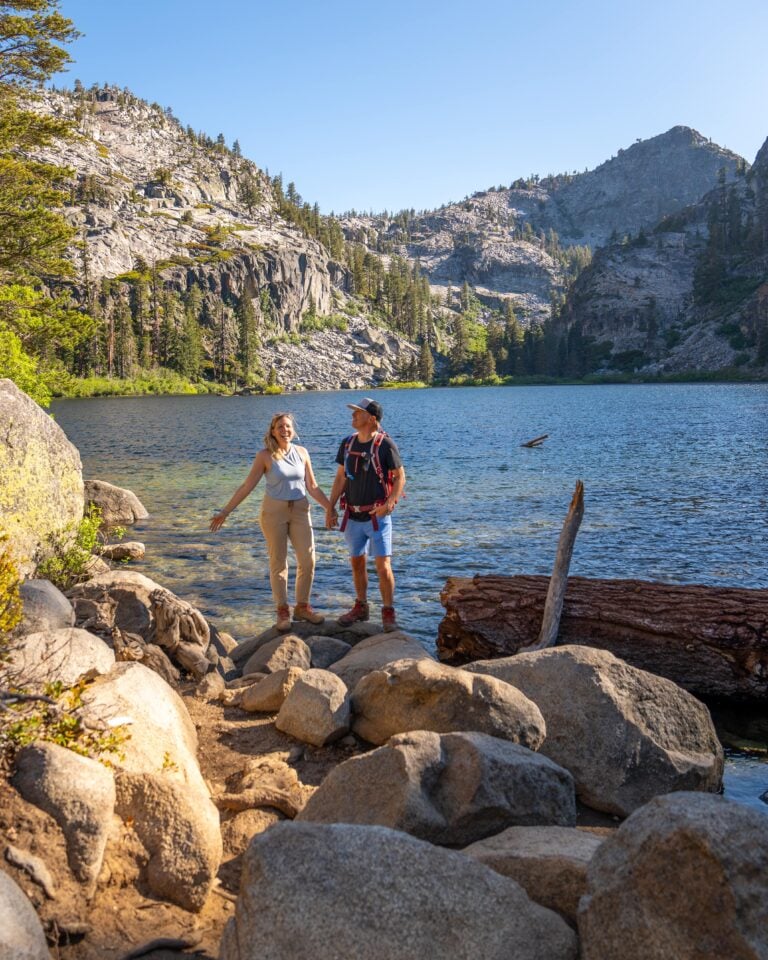 A couple stands on rocky ground by a clear mountain lake, surrounded by pine trees and rugged cliffs, holding hands and smiling under a bright blue sky.