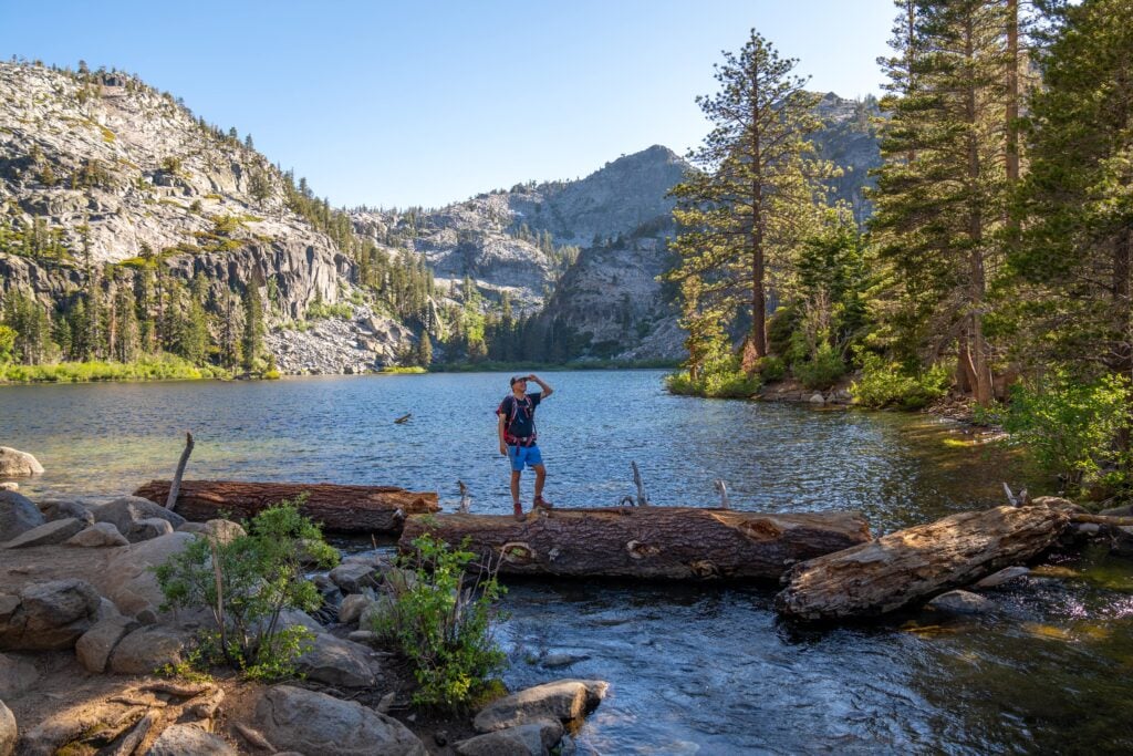 A person wearing a hat and backpack stands on a log by a clear mountain lake, surrounded by rocks, trees, and rugged cliffs under a bright blue sky.
