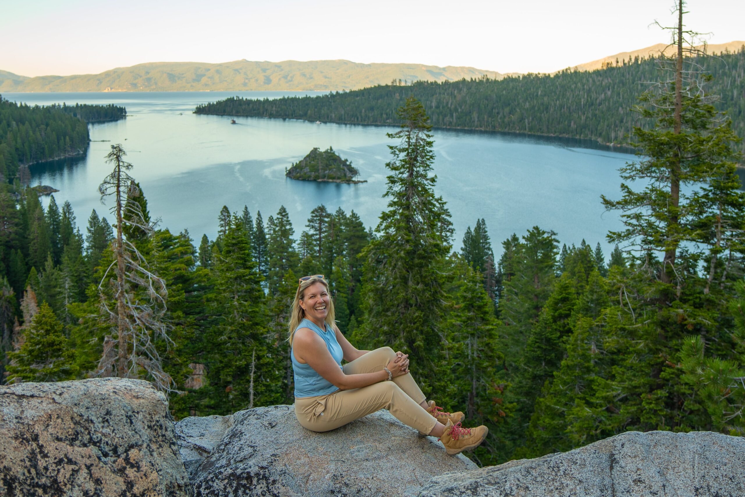 A woman sits smiling on a rock overlooking a scenic lake surrounded by pine trees, with an island in the water and forested hills in the background under a clear sky.