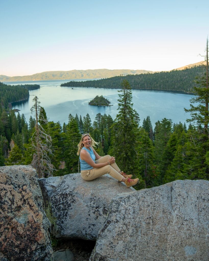 A woman sits on a large rock overlooking a scenic lake surrounded by pine trees and mountains under a clear sky. An island is visible in the middle of the lake.