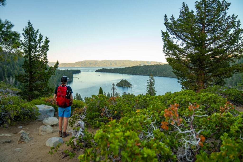 A hiker with a red backpack stands on a rocky trail, overlooking a scenic lake surrounded by pine trees and mountains under a clear sky. An island is visible in the middle of the lake.