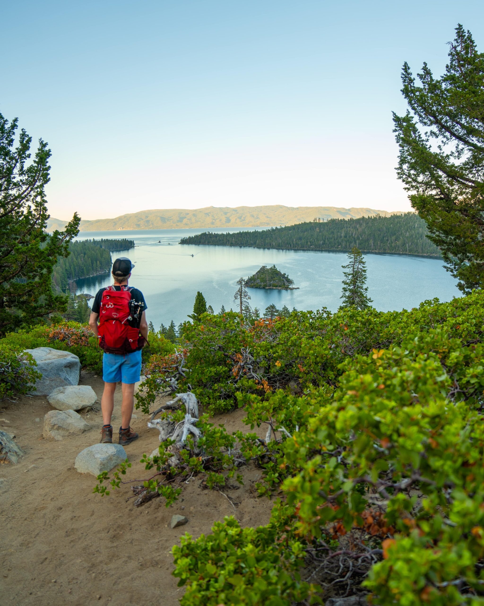 A person with a red backpack stands on a dirt trail overlooking a forested lake with a small island, surrounded by green trees and distant hills under a clear sky.