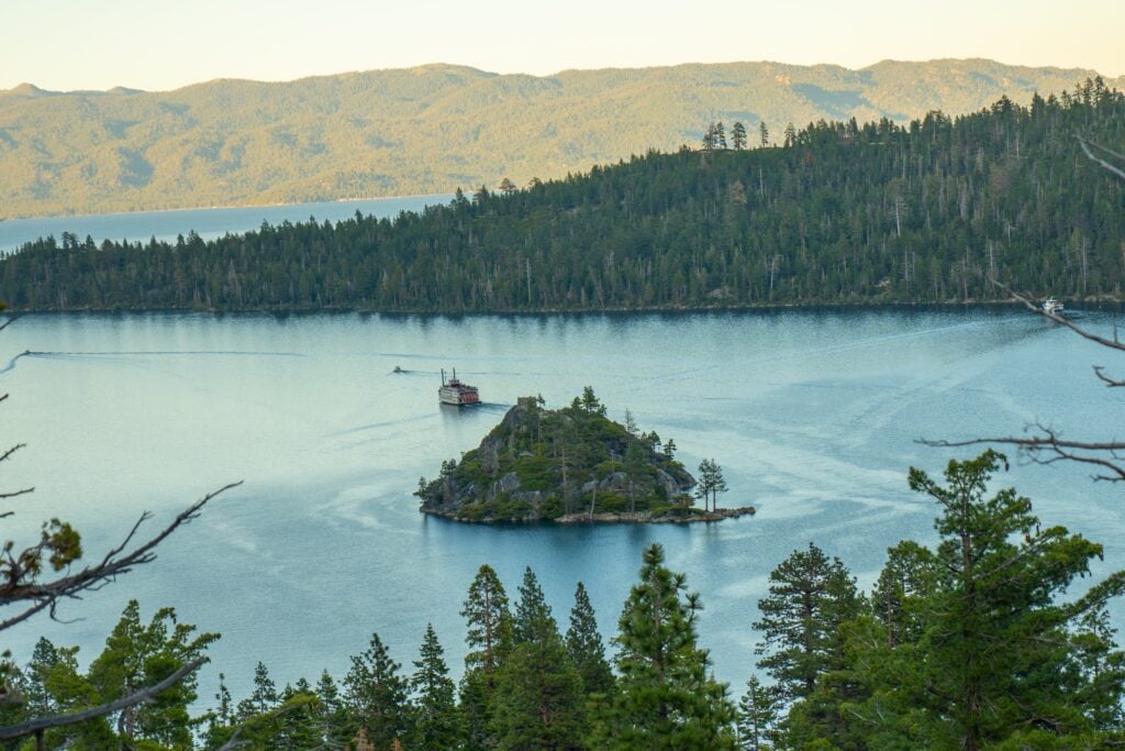 A scenic view of a small, tree-covered island in a calm lake surrounded by forested hills and distant mountains under a clear sky. A boat is visible near the island, and pine trees frame the foreground.