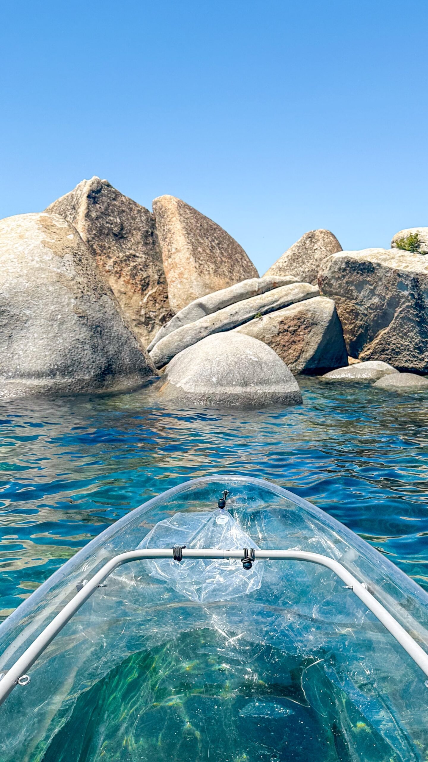 A clear kayak glides over vibrant blue water toward large, smooth boulders under a bright blue sky. The kayak’s transparent body reveals the water and rocks beneath.