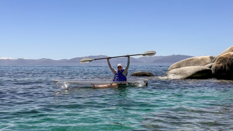 A person in a blue life vest sits in a clear kayak on turquoise water, holding a paddle above their head, near large rocks with mountains visible in the background under a clear blue sky.