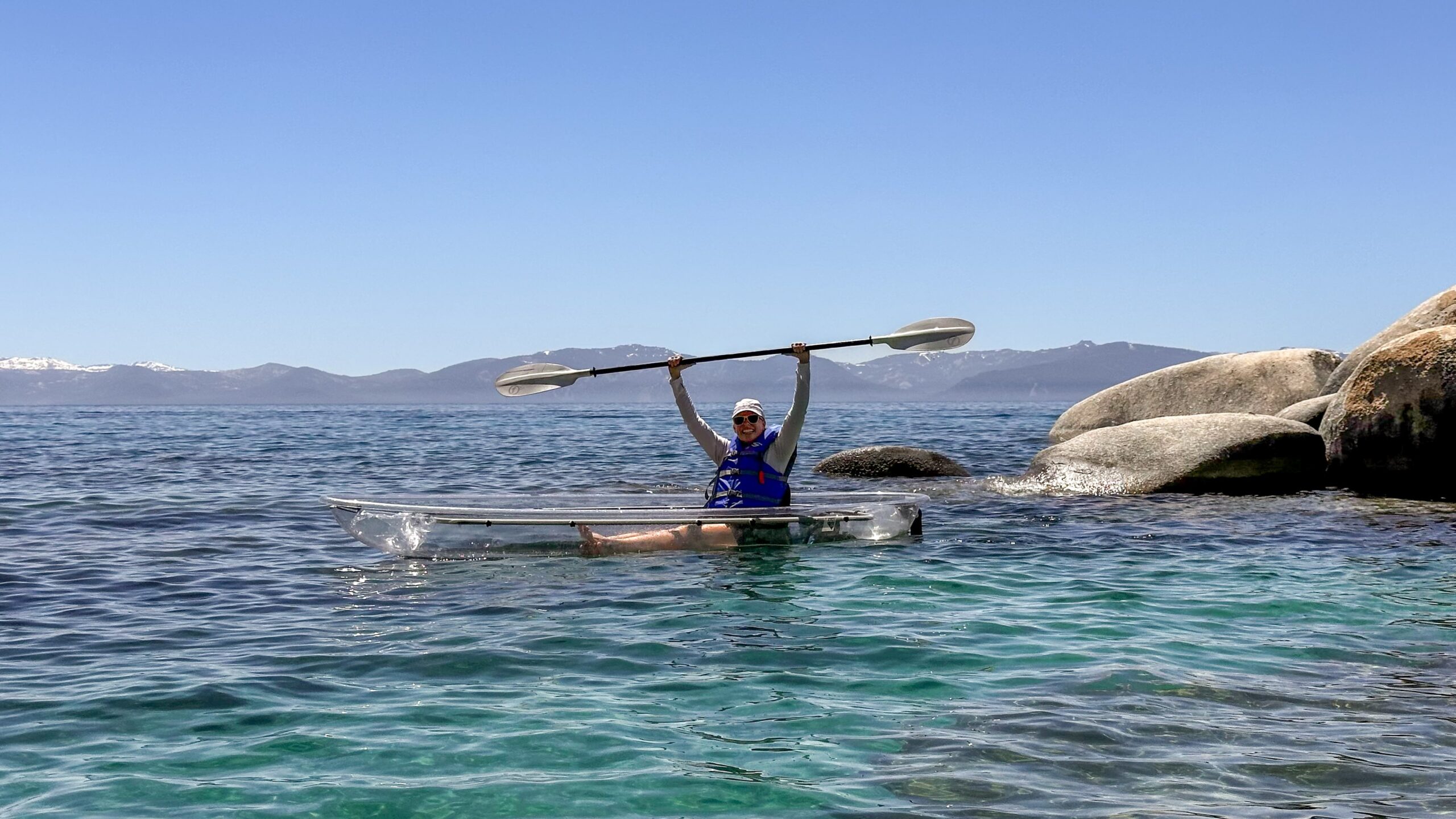 A person in a blue life vest sits in a clear kayak on turquoise water, holding a paddle above their head, near large rocks with mountains visible in the background under a clear blue sky.