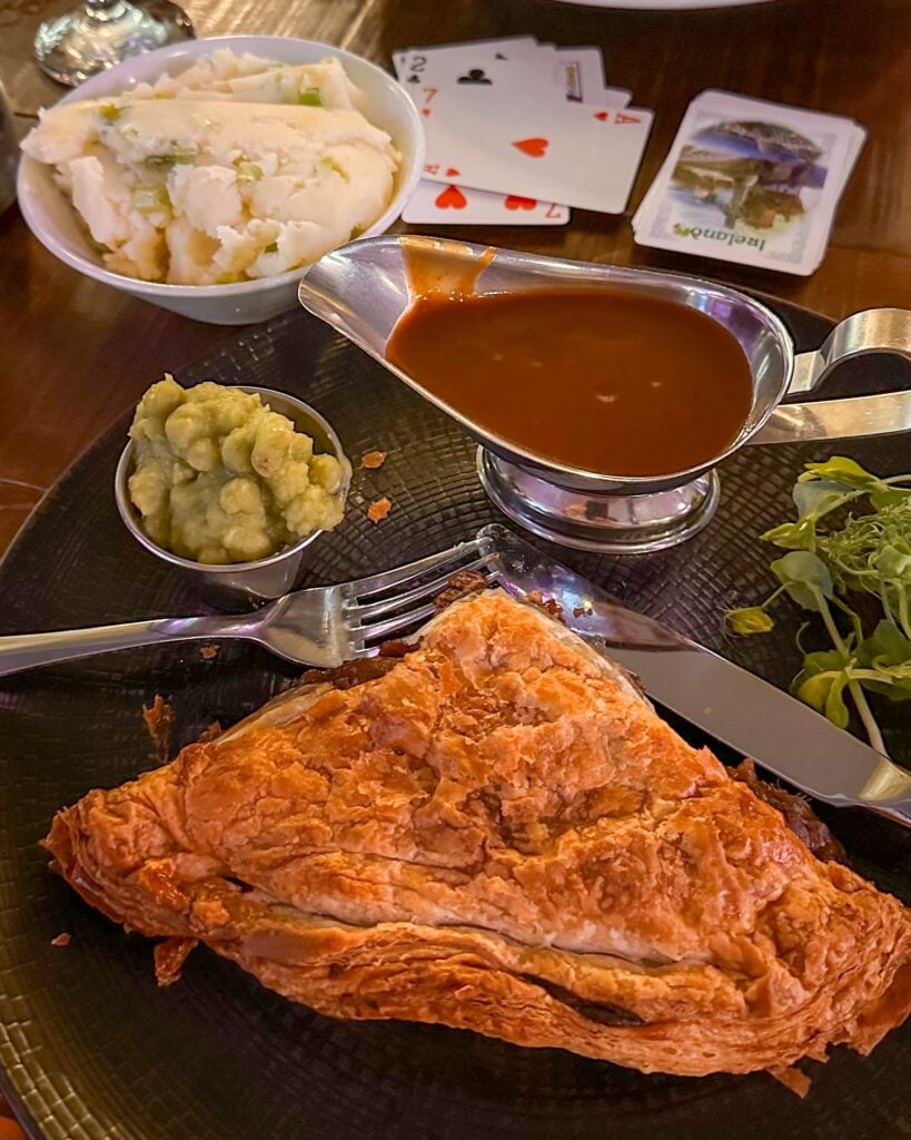 A plate featuring Irish food: a golden puff pastry pasty, mushy peas, gravy in a silver boat, mashed potatoes in a bowl, and a small salad. Playing cards are visible in the background.