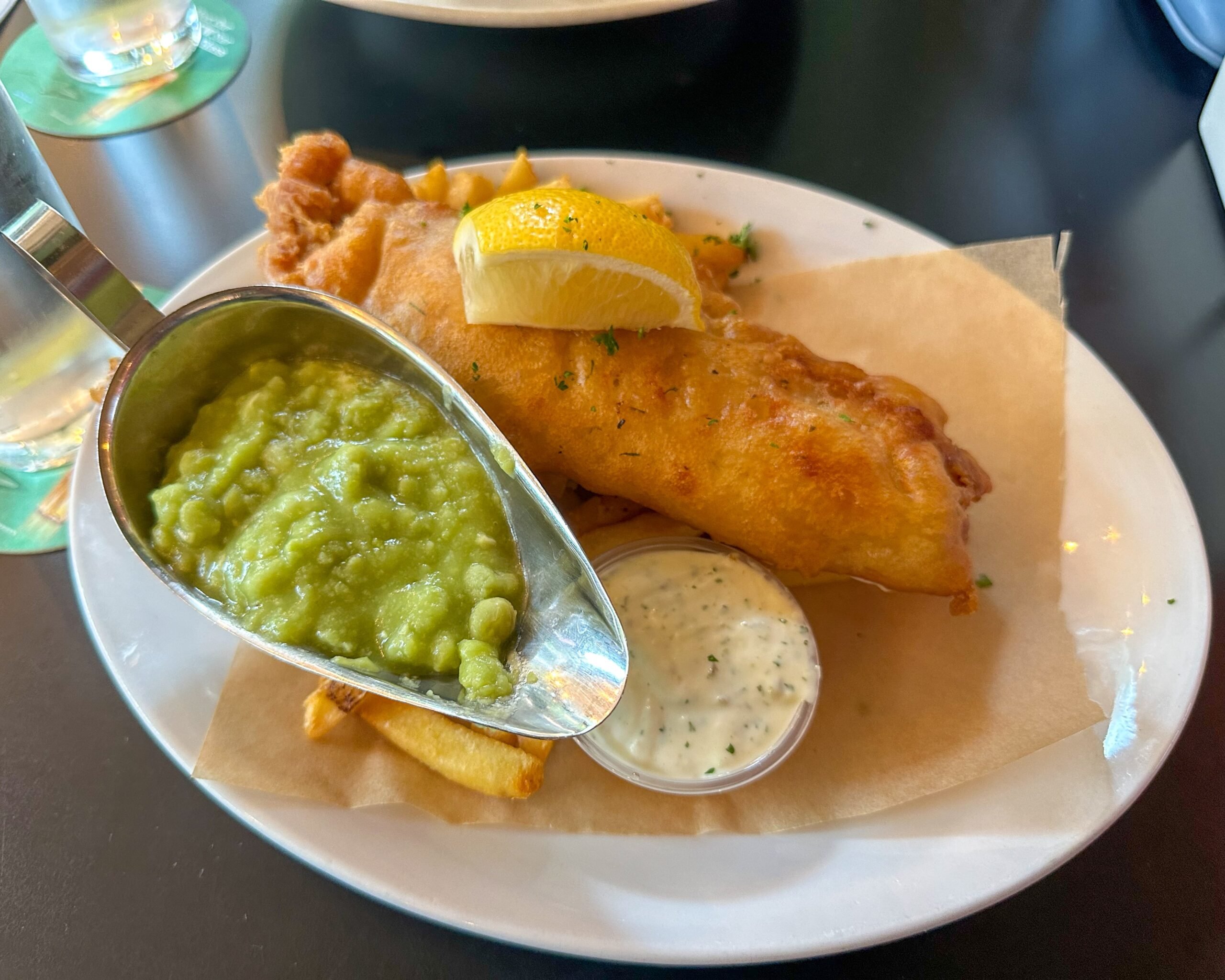 A classic plate of fish and chips, a staple of Irish cuisine, is served with a lemon wedge, tartar sauce, and a scoop of mushy peas in a metal gravy boat on parchment paper over a white plate.