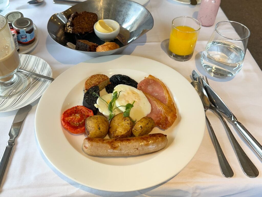 A plate of traditional English breakfast with sausage, bacon, fried egg, roasted potatoes, tomato, black pudding, and hash brown—perfect for those exploring Irish food or keen on Food in Ireland. Orange juice, bread with butter, and cutlery complete the setting.