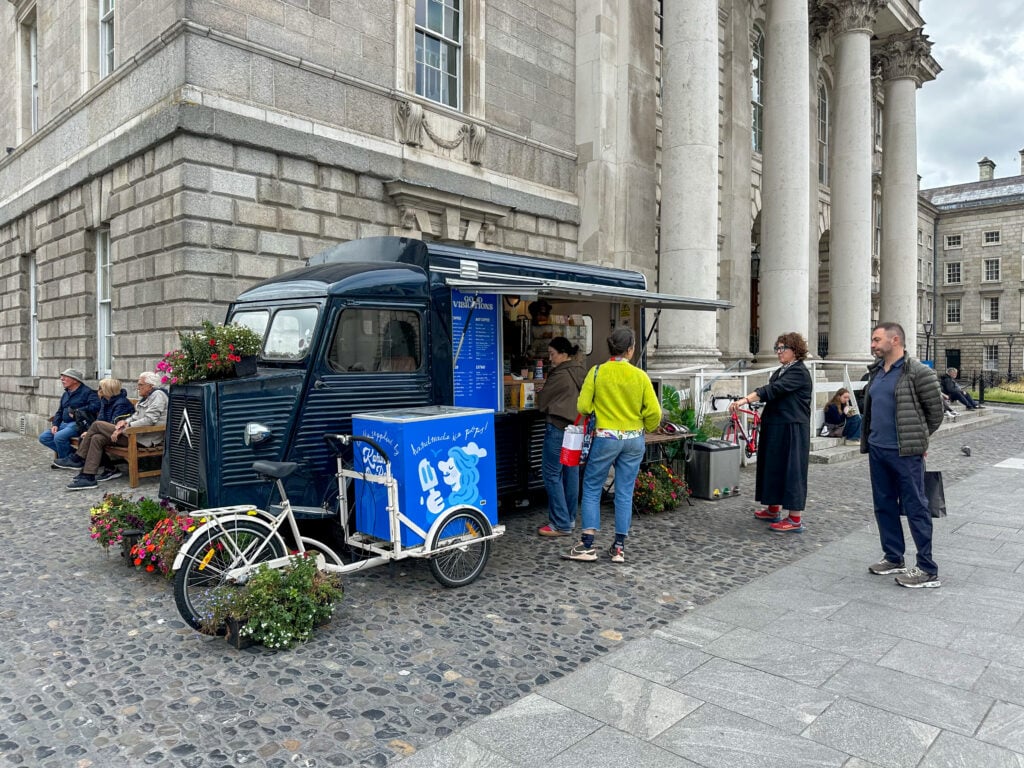 A small blue and black food truck is parked by a stone building. Several people stand in line to order, while others sit nearby. A decorated bicycle with flowers stands next to the truck.