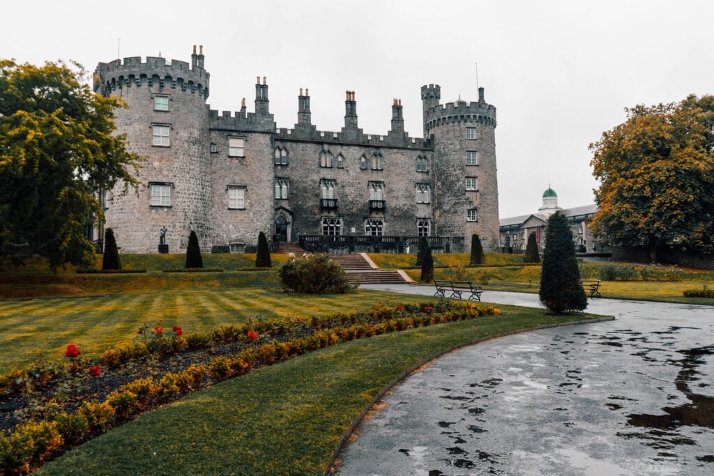A large stone castle with round towers and tall chimneys stands behind manicured lawns and flower beds, with a winding path and trimmed trees in the foreground on a cloudy day.