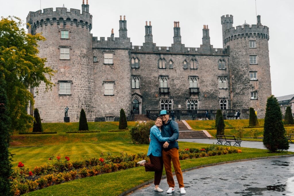 A smiling couple poses, hugging in front of a large stone castle with round towers and manicured gardens on a cloudy day. One person lifts a foot playfully, and both appear happy and relaxed.
