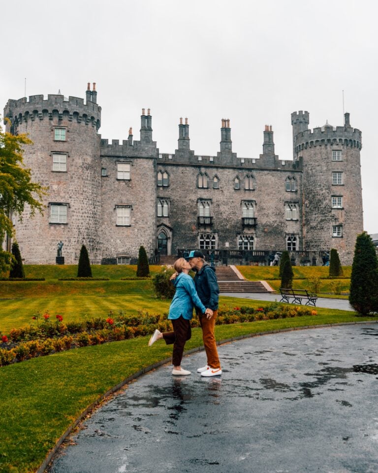 A couple shares a kiss on a wet pathway in front of a large stone castle with round towers and manicured gardens on a cloudy day.