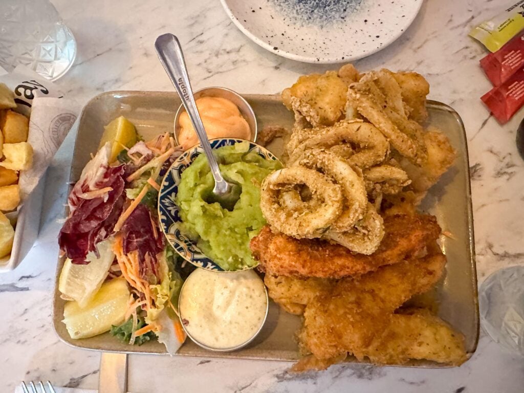 A plate of battered and fried seafood with dipping sauces, served with a side salad, lemon wedges, and mushy peas on a marble table—a classic Irish food and a must-try for anyone interested in Irish eating.