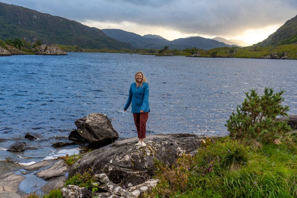 A woman stands on a rock by a lake surrounded by greenery and hills, under a cloudy sky with sunlight breaking through in the background.