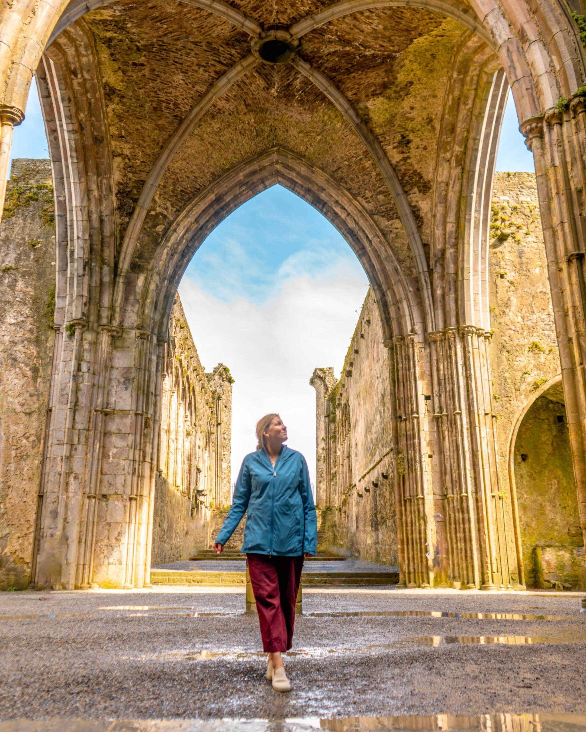 A person in a blue jacket and red pants walks through the ruins of an old stone cathedral with tall arches and sunlight streaming in.