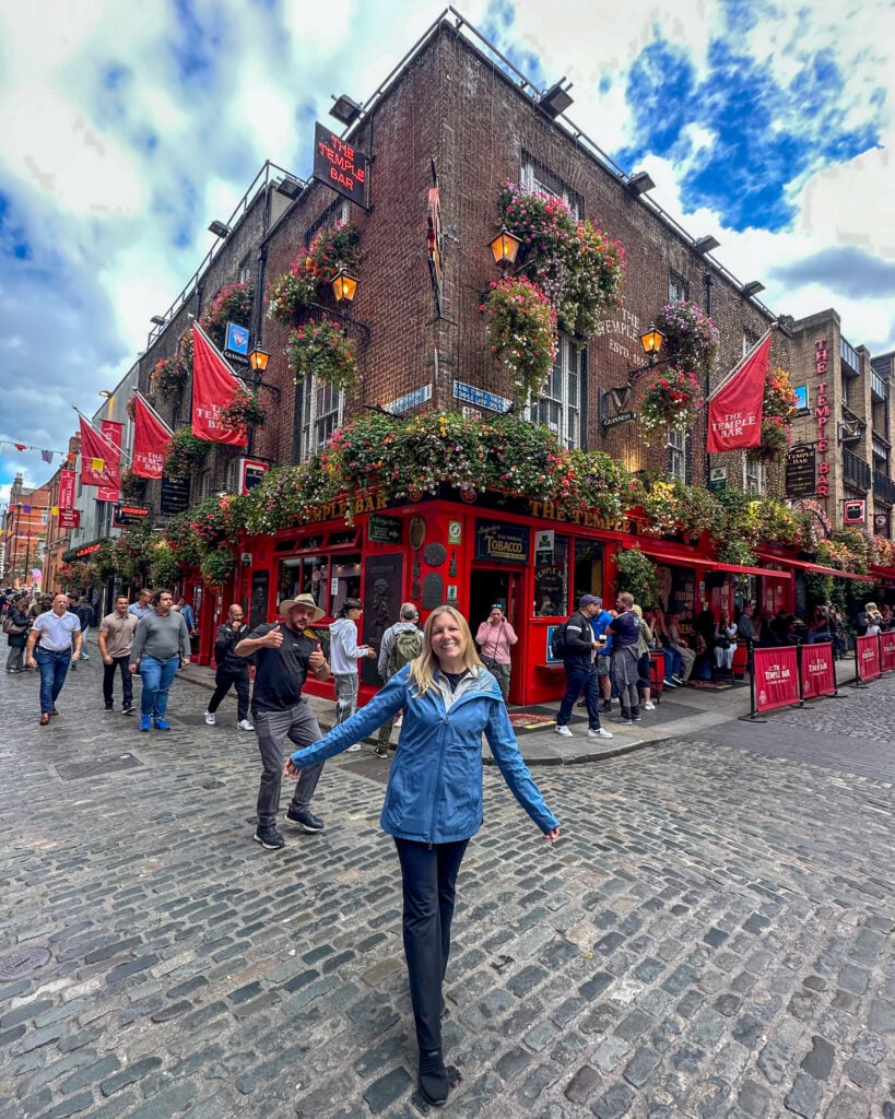 A woman in a blue jacket stands smiling with arms outstretched on a cobblestone street in front of a lively, flower-adorned pub with red trim, as people walk nearby under a partly cloudy sky.