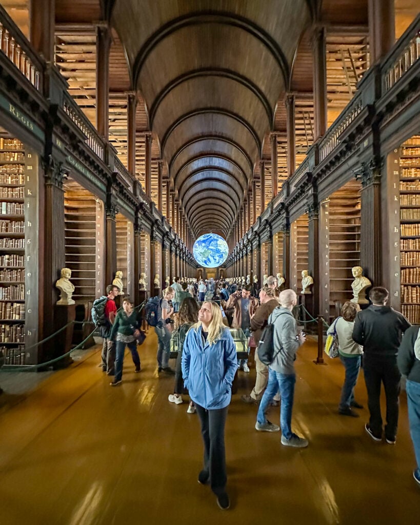 A group of people explore a grand, arched library hall lined with tall bookshelves and busts; a large illuminated globe hangs at the far end of the hall.