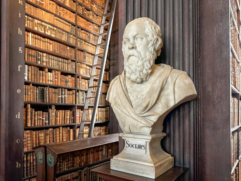 A marble bust labeled "Socrates" stands in front of dark wooden bookshelves filled with old books and a tall library ladder in a historic library setting.