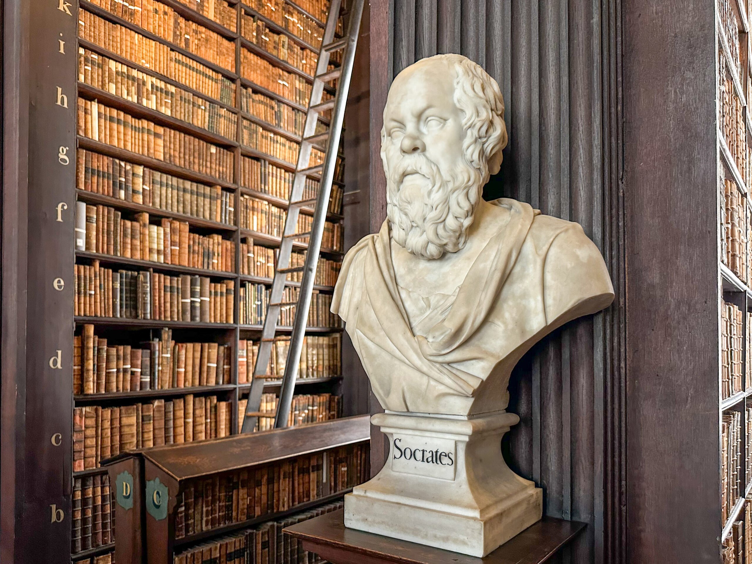 A marble bust labeled "Socrates" stands in front of dark wooden bookshelves filled with old books and a tall library ladder in a historic library setting.