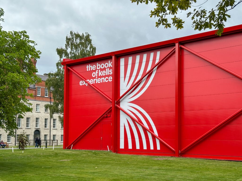 A large red building with bold white text reading "the book of kells experience" and a stylized graphic, set on a grassy area with trees and a brick building in the background.