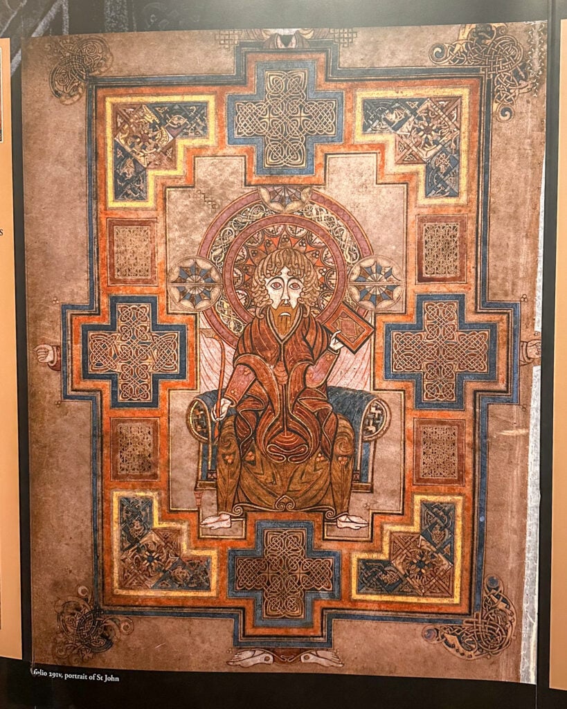 Illuminated medieval manuscript page featuring a seated, haloed figure holding a book, surrounded by ornate geometric patterns, intricate knotwork, and decorative crosses. Text in lower left reads "folio 291v, portrait of St John.