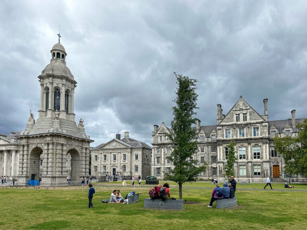 People sit and relax on a grassy lawn in front of historic stone buildings, including a domed bell tower, under a cloudy sky at a university campus.